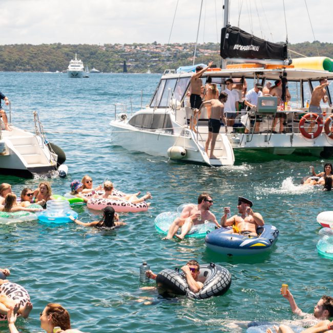 People are reveling in a sunny day, floating on inflatable rafts and swimming near two anchored catamarans during a lively Catamaran party Sydney.