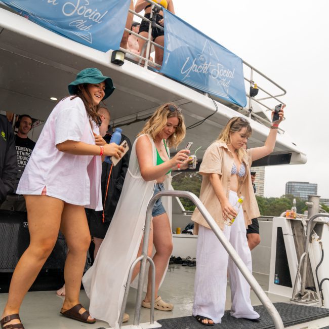 People are dancing and enjoying a party on a yacht. Some are on the upper deck, and others near the entrance with drinks in hand. The yacht has "The Yacht Social Club" banners fluttering as the Catamaran party Sydney vibes fill the air.