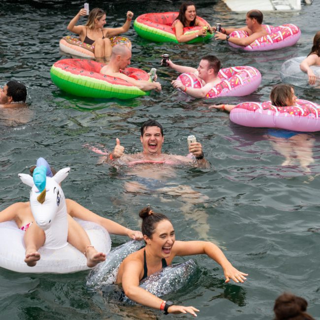People floating on inflatables in a body of water. They appear to be having fun, with some holding drinks and others swimming. Brightly colored inflatables include swans, unicorns, and donuts—a scene reminiscent of a Sydney boat party hire or luxury yacht hire Sydney adventure.