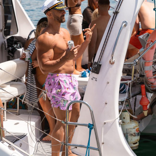 A man wearing pink shorts and a captain's hat holds a drink and points while standing on a crowded boat, enjoying a catamaran party on Sydney Harbour.