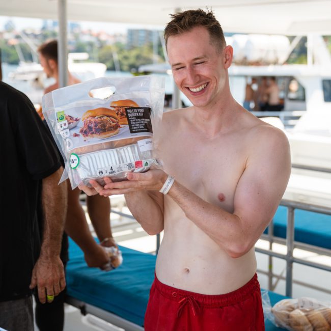 A smiling man with short hair is holding a pack of Hawaiian rolls on a boat, shirtless and wearing red shorts. Other passengers and the sea are in the background, enjoying the Catamaran party Sydney vibes.
