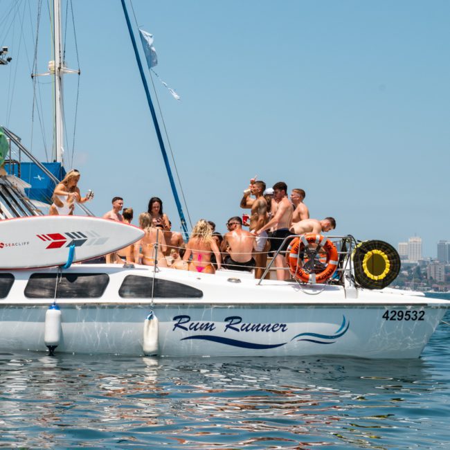 A group of people relaxes on a catamaran named "Rum Runner," anchored on calm waters with city buildings visible in the background, enjoying a lively Sydney boat party hire.