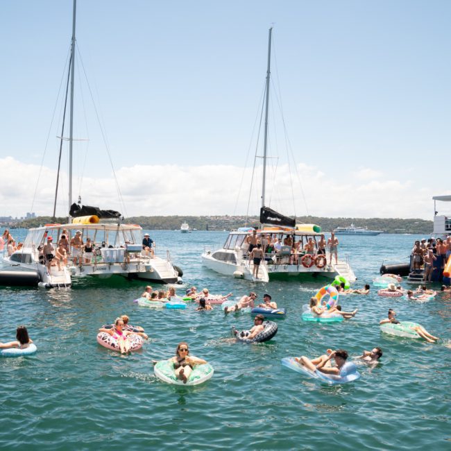 People on boats and inflatables are gathered on a sunny day in a calm body of water, enjoying leisure activities. It's the perfect setting for a private yacht charter on Sydney Harbour, making the experience even more memorable.