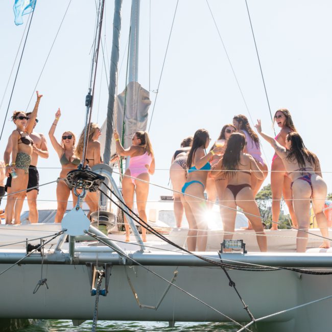 A group of people in swimwear are standing and socializing on a luxury yacht hire Sydney catamaran on a bright, sunny day.
