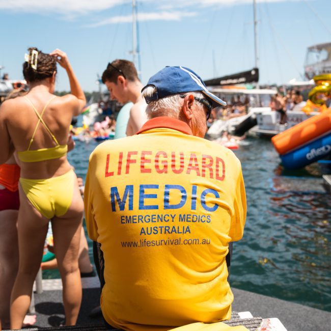A lifeguard medic in a yellow shirt sits by the water, with several people and boats visible in the background on a sunny day. The scene is perfect for Corporate boat events Sydney or even a Private yacht charter Sydney Harbour.