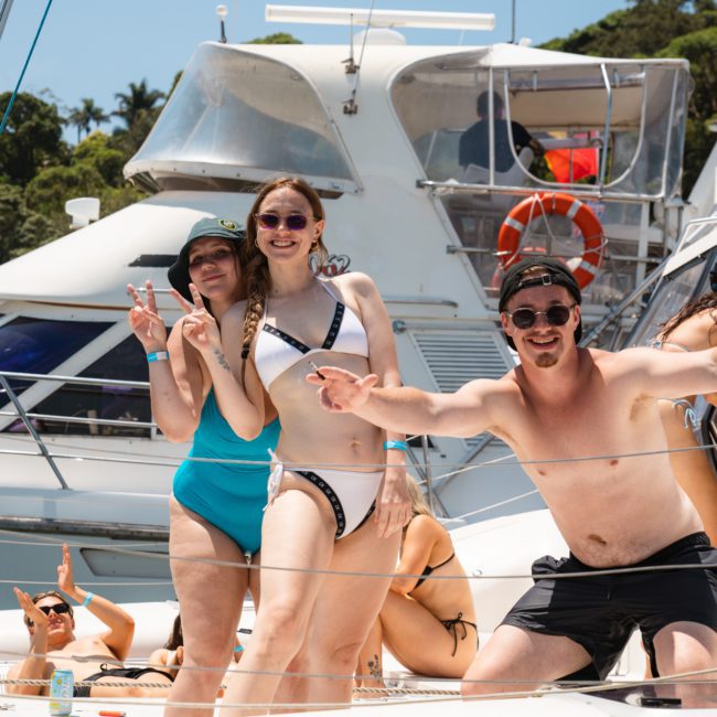 Three adults in swimwear smiling and posing on a boat, with two women in the background lounging. Boats and trees are visible in the background on a sunny day, reflecting the fun of a Catamaran party Sydney.