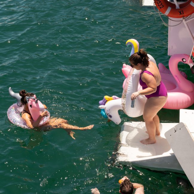 People floating on inflatable toys in a body of water near a private yacht charter in Sydney Harbour. One person is on a unicorn float, another on a flamingo, and two others are swimming nearby.