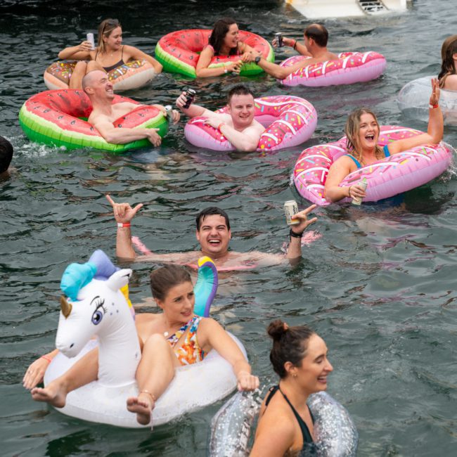 People floating in water on inflatable pool toys, including a unicorn, during a Sydney boat party hire. Some are holding drinks and smiling or making hand gestures.