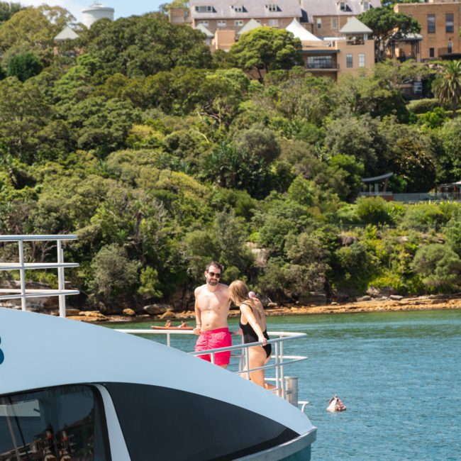Two people stand on the deck of a luxury yacht hired in Sydney Harbour, docked near a lush green shoreline with buildings in the background.