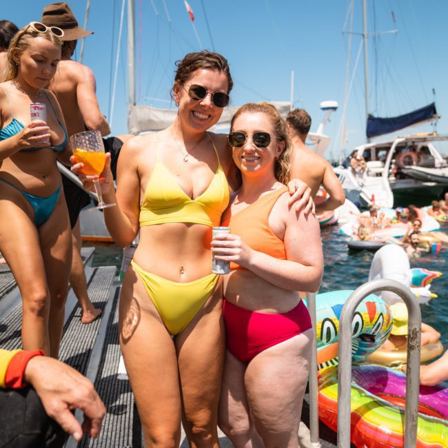 Two people in swimsuits stand on a dock with drinks in hand, surrounded by others in swimwear near a pool and boats on a sunny day, enjoying the Catamaran party Sydney.
