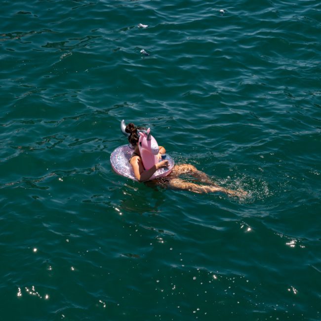 A person floats on a pink inflatable ring in a body of water, wearing a hat and holding a drink, enjoying the view from their luxury yacht hire Sydney.