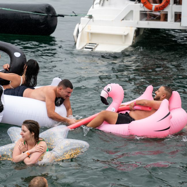 Adults swim and relax on inflatable pool floats, including a black swan and a pink flamingo, near a boat named "Rum Runner," perfect for a Catamaran party Sydney.