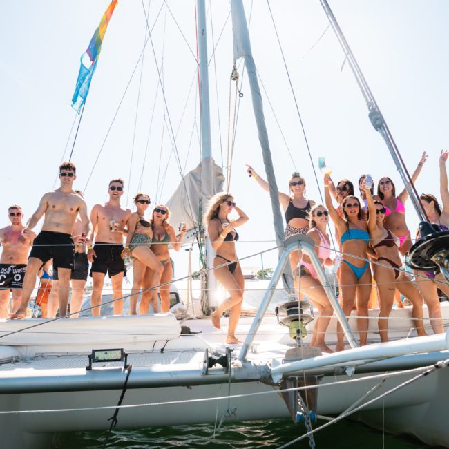 A group of people in swimsuits enjoy their time on the deck of a sailing boat, with some posing and others raising their hands. The weather is sunny, and a colorful flag is visible in the background, epitomizing the perfect setting for a Sydney boat party hire.