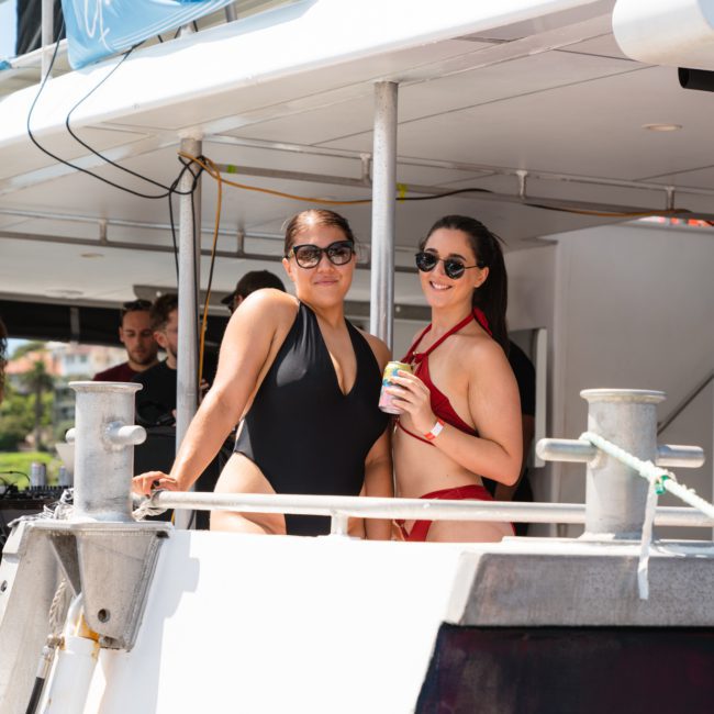 Two women in swimsuits stand on a boat deck, smiling and wearing sunglasses. One holds a drink can. They look ready for an unforgettable catamaran party in Sydney.