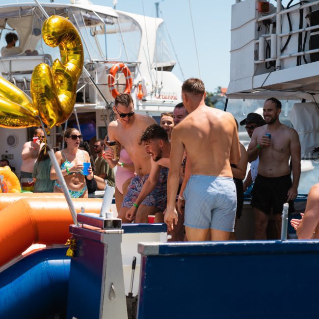 A group of people in swimwear are gathered on a luxury yacht hire in Sydney, celebrating with drinks and a large gold balloon shaped like the number "30." Some are standing on the yacht, while others are near an inflatable slide.