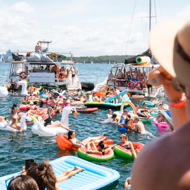 A lively scene with people on inflatable floats and boats celebrating in a harbor under a sunny sky, with a person in a hat observing in the foreground. Perfect for Sydney boat party hire!