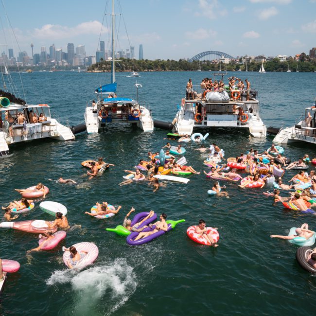 A bustling scene of people on boats and inflatable pool toys partying on the water with a city skyline and bridge visible in the background, featuring a lively catamaran party in Sydney.
