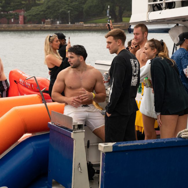 A group of people is gathered on a boat. A man wearing white shorts stands at the edge while others chat and look on. The background shows trees and water. An inflatable slide is visible behind the man, perfect for a Catamaran party Sydney adventure.