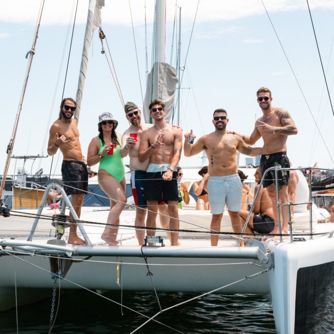 A group of five people stands on the deck of a catamaran, posing and smiling at the camera. The sunny day features a blue sky and calm water in the background, perfect for a private yacht charter on Sydney Harbour.