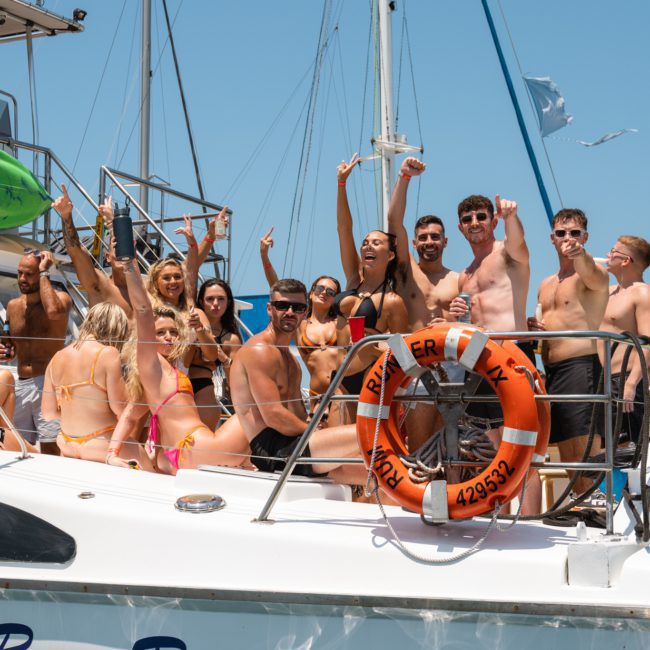 A group of people in swimwear enjoying a sunny day on a luxury yacht hire Sydney. They are smiling, raising their arms, and some are posing for the camera. The yacht is equipped with life-saving gear and kayaks on top, perfect for a lively catamaran party Sydney.