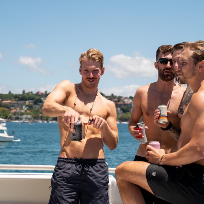 Four men in swim trunks are enjoying drinks on a luxury yacht hire in Sydney. The background features a clear blue sky, a body of water, and another boat in the distance.