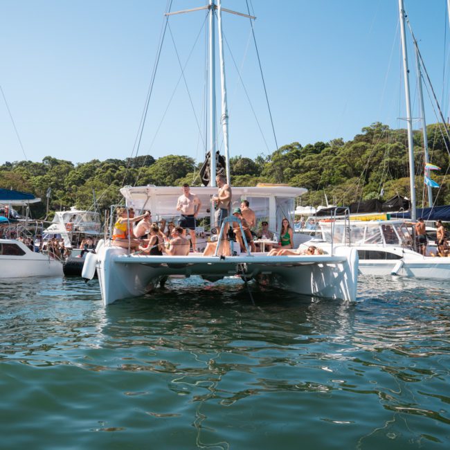 A group of people relaxing and socializing on a luxurious catamaran sailboat, surrounded by other boats on a sunny day with a forested shoreline in the background, enjoying the ambiance of their private yacht charter Sydney Harbour.