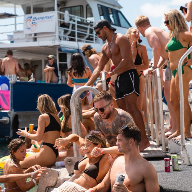 A group of people in swimwear enjoying a pool party on a docked boat. Some are sitting, standing, or lounging on inflatables in the water, while others are socializing on the boat. Perfect for those looking to experience DJ boat hire Sydney for any occasion.