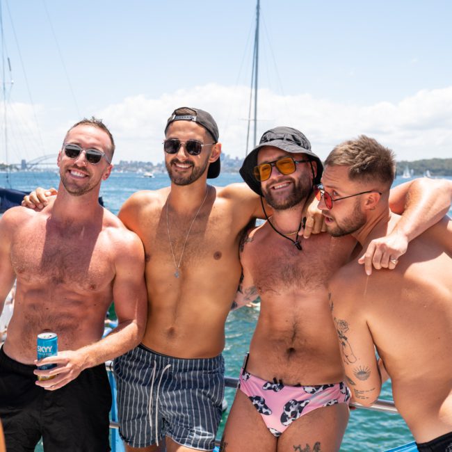 Four men in swimwear pose together on a luxury yacht with a body of water and other boats in the background, enjoying what appears to be an exclusive catamaran party in Sydney.