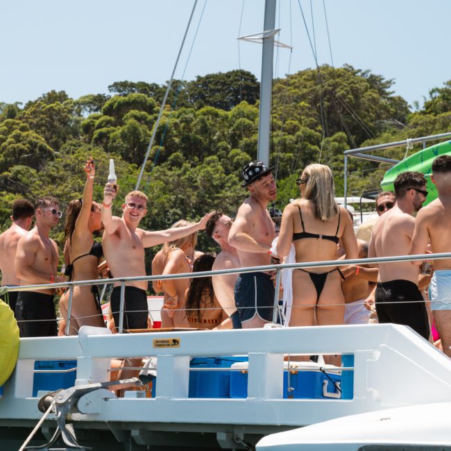 A group of people in swimwear are standing and socializing on a boat near a forested shoreline. Some are holding drinks, and the atmosphere appears lively and festive. It looks like an unforgettable DJ boat hire Sydney event, perfect for summer fun.