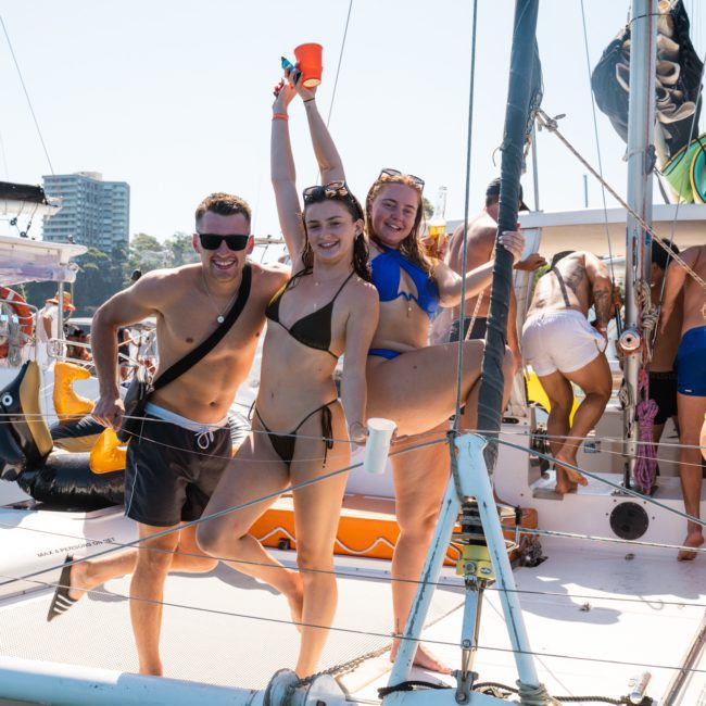 A group of people wearing swimwear are enjoying themselves on a private yacht charter in Sydney Harbour under sunny skies. Some are posing and smiling for the camera, while others are mingling or relaxing near the boat's railing.