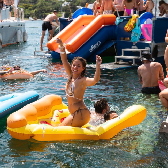 Several women and men in swimsuits enjoy a sunny day on the water with inflatable toys, slides, and boats. One woman in the foreground sits on a yellow inflatable and smiles at the camera, capturing the joy of a Sydney boat party hire.