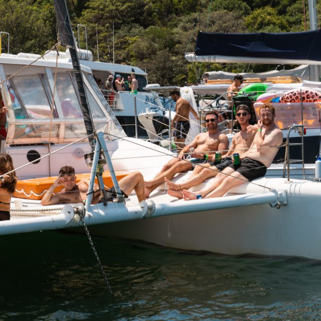A group of people in swimwear are lounging on the deck of a white catamaran boat, with other boats and greenery visible in the background, enjoying their Sydney boat party hire.