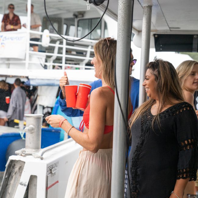 People socializing on a dock beside a boat, some holding red cups, while others engage in conversation. Enjoy the vibrant atmosphere of this DJ boat hire Sydney experience.