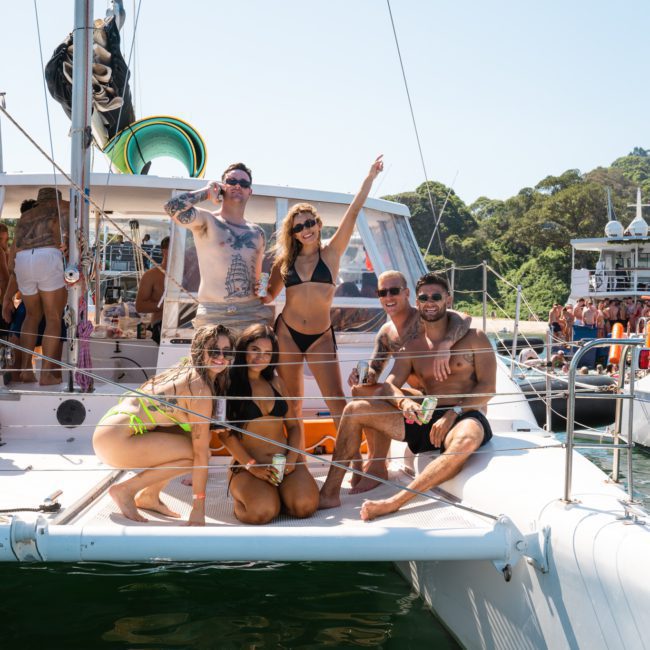 A group of people in swimwear are posing and smiling on the deck of a boat anchored in the water. Other boats and people are visible in the background, creating a vibrant atmosphere perfect for a catamaran party Sydney event.