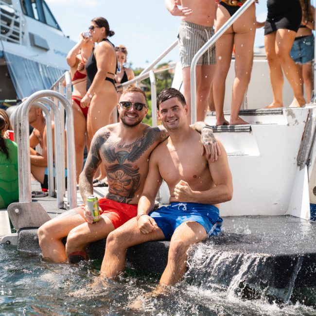 Two people in swimwear sit on the edge of a boat, smiling, with others in swimwear around. One person has a tattoo and holds a drink. Water splashes near them, capturing the excitement of a luxury yacht hire Sydney experience.
