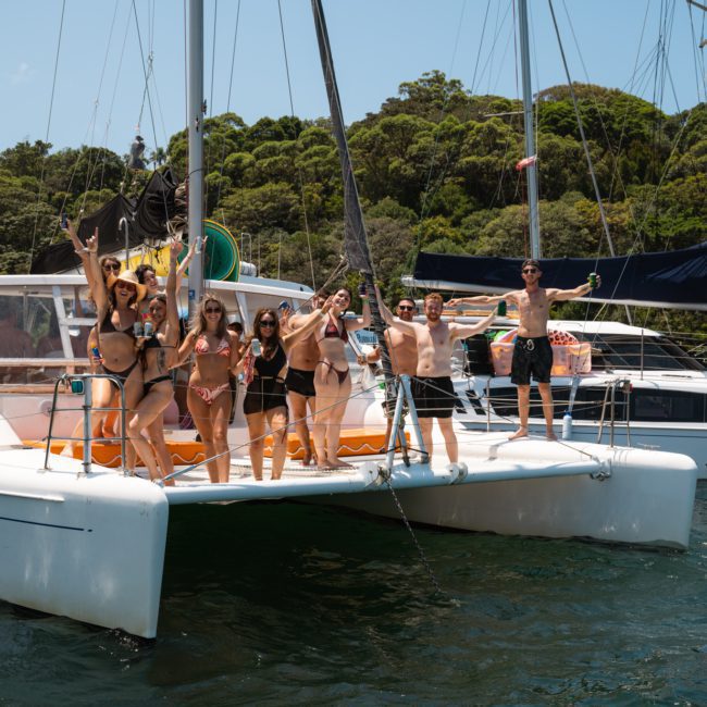 A group of people in swimwear pose and celebrate on the deck of a catamaran yacht on a sunny day with a forested shore in the background, enjoying their private yacht charter Sydney Harbour.