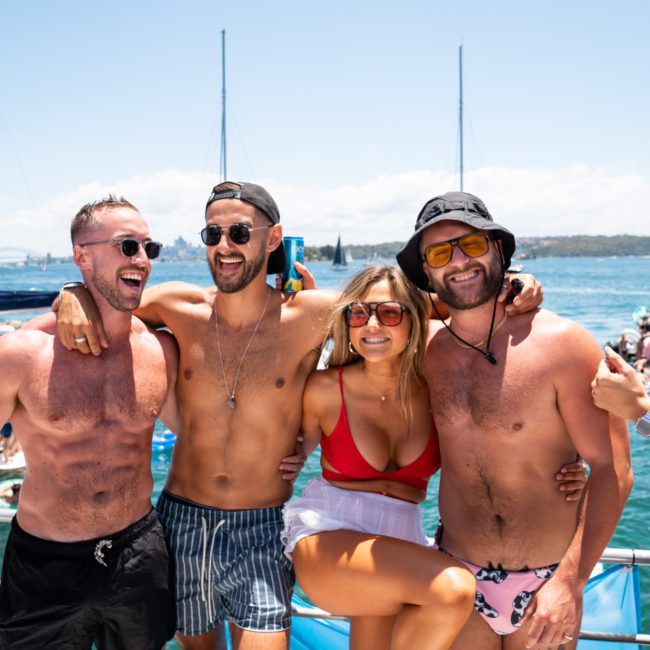 Four friends, three shirtless men and a woman in a red bikini top, smile and pose together on a catamaran party in Sydney Harbour with other boats and water activities in the background.
