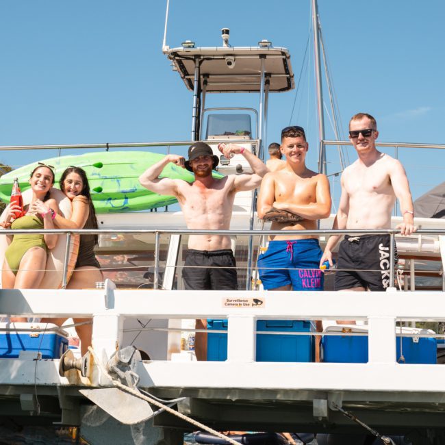 A group of five people pose on the deck of a boat on the water, with kayaks and blue containers visible behind them, under a clear sky. Perfect for corporate boat events in Sydney or those looking for a private yacht charter on Sydney Harbour.