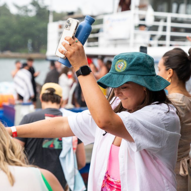 A woman in a green bucket hat and white shirt dances while holding a smartphone and a blue water bottle. People and a luxury yacht are in the background.