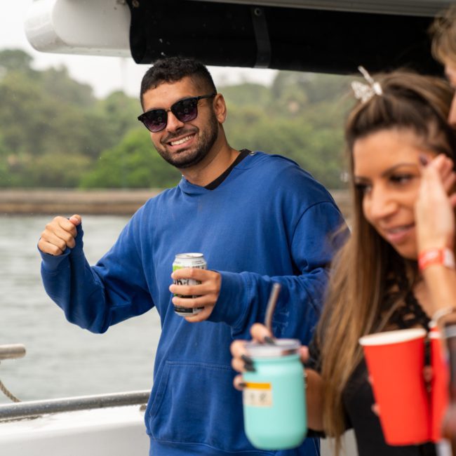 A person in sunglasses and a blue hoodie smiles while holding a can, standing on a boat during a Sydney boat party hire. Another person in the foreground holds a cup and looks away. Trees and water are visible in the background, creating the perfect setting for this private yacht charter Sydney Harbour experience.