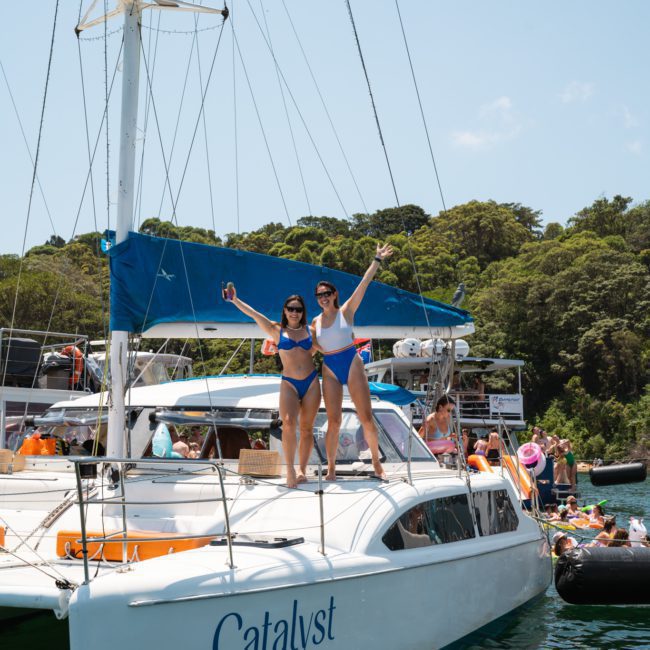 Two women in blue swimwear stand on the deck of a sailboat named Catalyst, posing with raised arms. Other boats and people can be seen in the water and on a dock in the background, enjoying their private yacht charter Sydney Harbour experience.