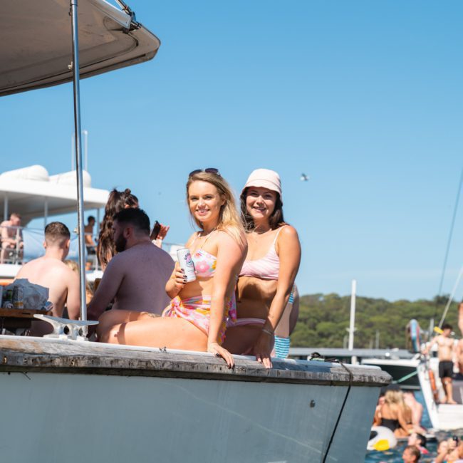 Two women in swimsuits sit on the edge of a luxury yacht, smiling at the camera. Several people are on boats in the background, enjoying a sunny day on the water.