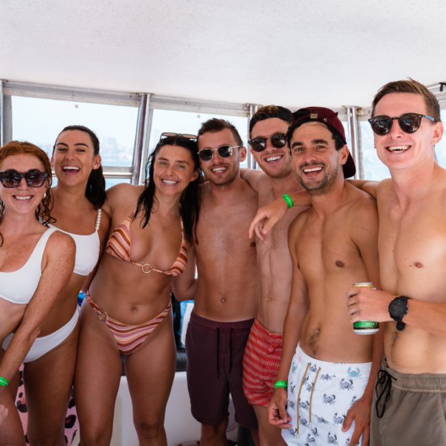 A group of seven people in swimwear, smiling and posing for a photo on a private yacht charter in Sydney Harbour. Sunlight streams through the windows, and an ocean view is visible outside.