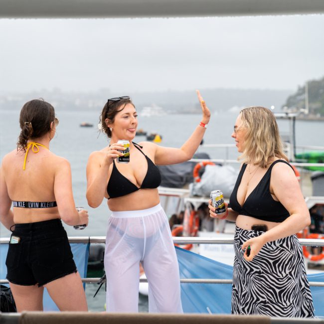 Three women in swimwear stand on a boat, holding drinks and interacting. The background shows a foggy waterfront with other boats visible. Experience the ultimate celebration with a private yacht charter Sydney Harbour.