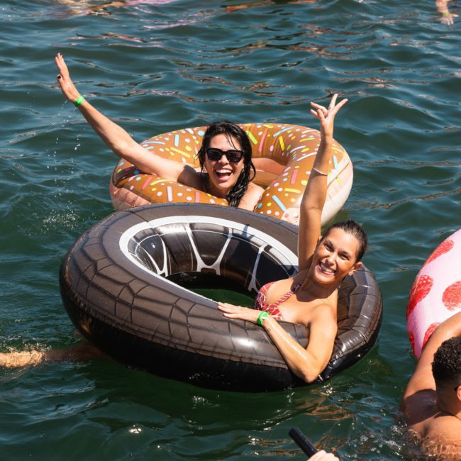 Two women in inflatable rings smile and raise their arms while floating on a body of water, surrounded by others in various inflatables during a vibrant catamaran party Sydney.