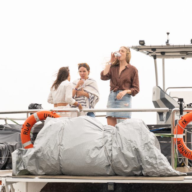 Three women stand on a boat, with two conversing while the third drinks from a glass. Two life rings are attached to the railing. The boat is outdoors and appears docked, ideal for a private yacht charter on Sydney Harbour or a Sydney boat party hire.