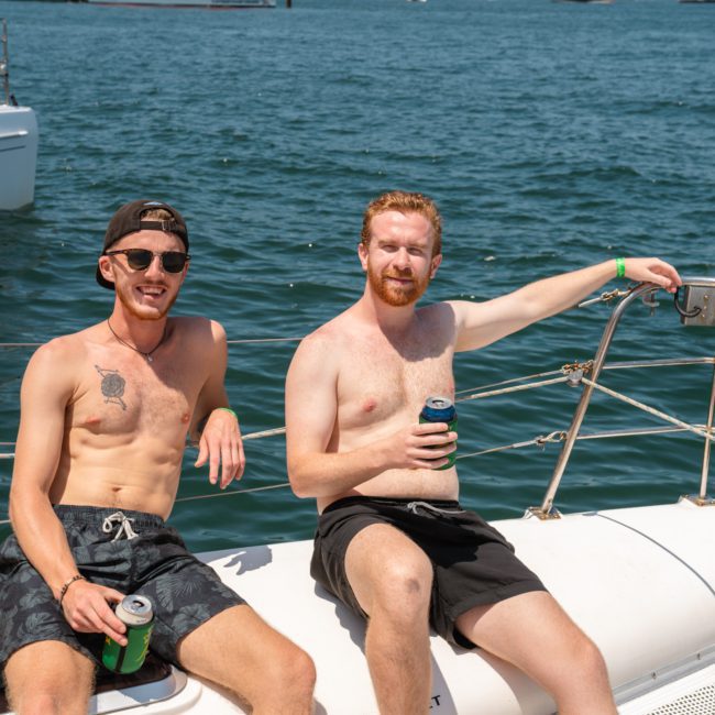 Two men sitting shirtless on a boat's bow, holding drinks. They are wearing sunglasses and swim shorts. The sea and another boat named "Pegasus" are visible in the background, embodying the essence of a luxury yacht hire Sydney experience.