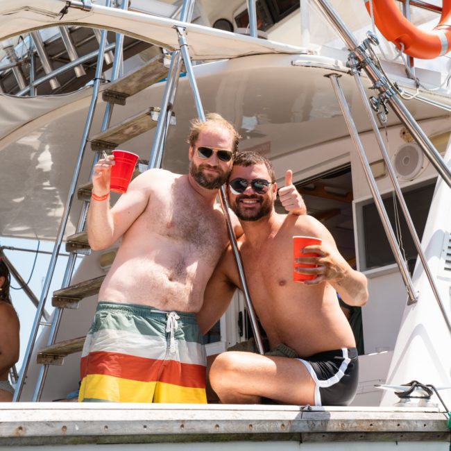 Two men in swim trunks, holding red cups, stand on a boat deck. One gives a thumbs-up, both smiling at the camera during a luxury yacht hire in Sydney.