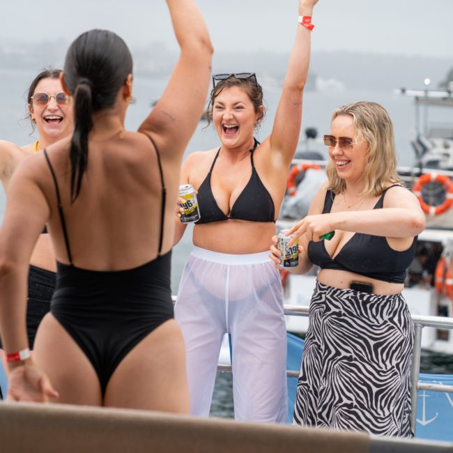 Four women in swimwear cheer and hold drinks on a boat, enjoying a Private yacht charter Sydney Harbour with a stunning waterfront backdrop.