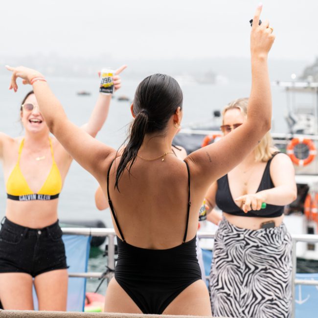 A group of people in swimsuits is dancing and celebrating on a luxury yacht hire Sydney. They are smiling and enjoying a sunny day on the water.